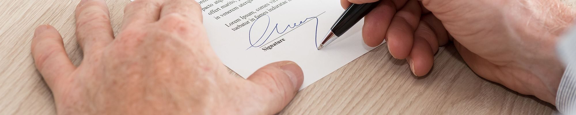 Deux mains en gros plan, la main droite tient un crayon et signe un document papier poser sur le bureau.