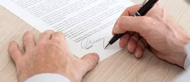 Deux mains en gros plan, la main droite tient un crayon et signe un document papier poser sur le bureau.