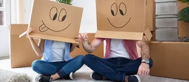 Une femme et un homme assis en tailleur dans une pièce avec des cartons, les deux personnes ont un carton sur la tête avec un dessin de visage souriant dessus.