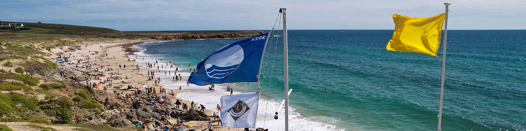Plage de Trez Goarem par beau temps avec de nombreux baigneurs, vue prise depuis les hauteurs, on voit le drapeau du label pavillon bleu.
