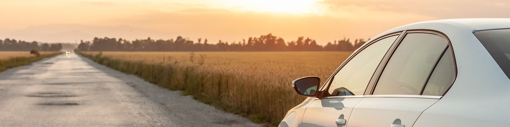 Une voiture blanche qui roule sur une route de campagne, le soleil couchant illumine la route de tons orangés.