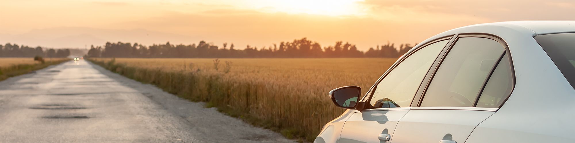 Une voiture blanche qui roule sur une route de campagne, le soleil couchant illumine la route de tons orangés.