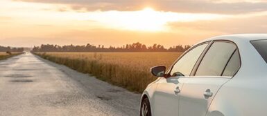 Une voiture blanche qui roule sur une route de campagne, le soleil couchant illumine la route de tons orangés.