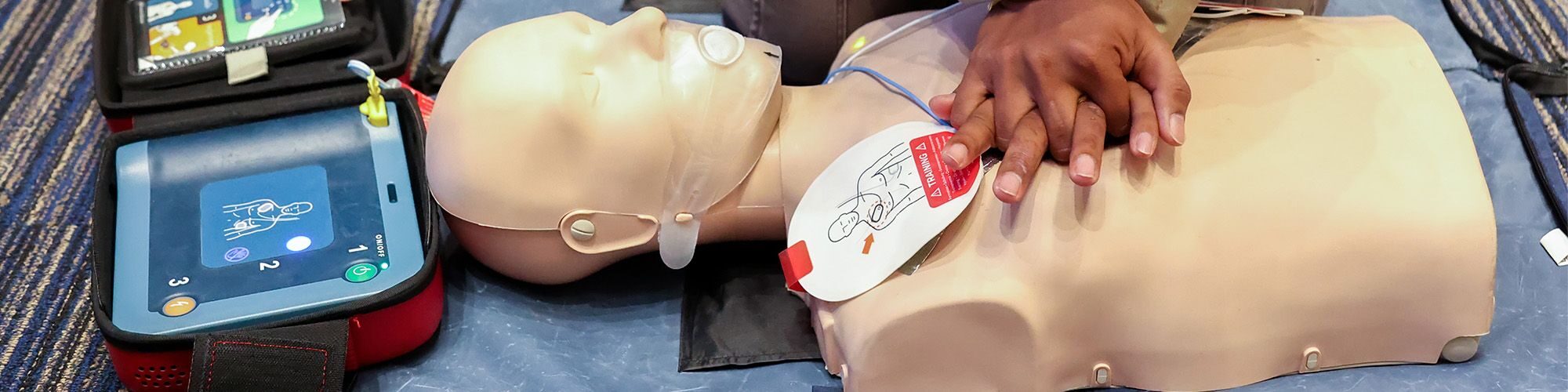 Une personne qui fait un massage cardiaque à un mannequin, , sur sa gauche il un défibrillateur qui est connecté au mannequin avec deux électrodes.