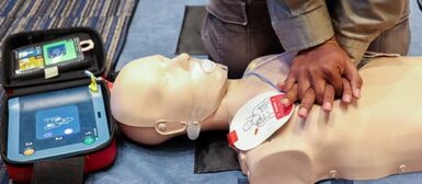 Une personne qui fait un massage cardiaque à un mannequin, , sur sa gauche il un défibrillateur qui est connecté au mannequin avec deux électrodes.
