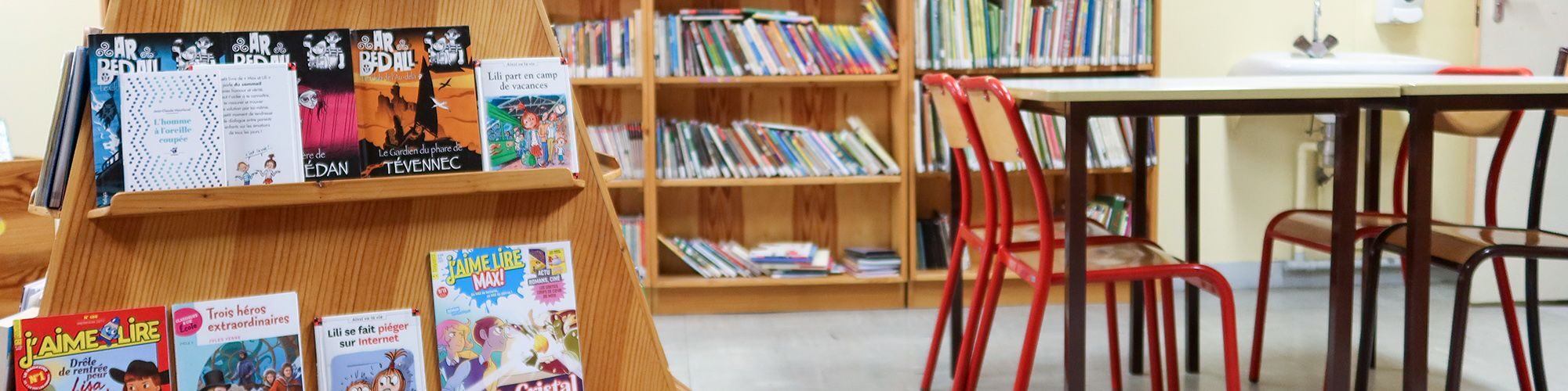 Salle de classe, avec une table et 4 chaises d'écoliers. Dans le fond, il y a des étagères avec des livres pour enfants.