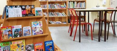 Salle de classe, avec une table et 4 chaises d'écoliers. Dans le fond, il y a des étagères avec des livres pour enfants.