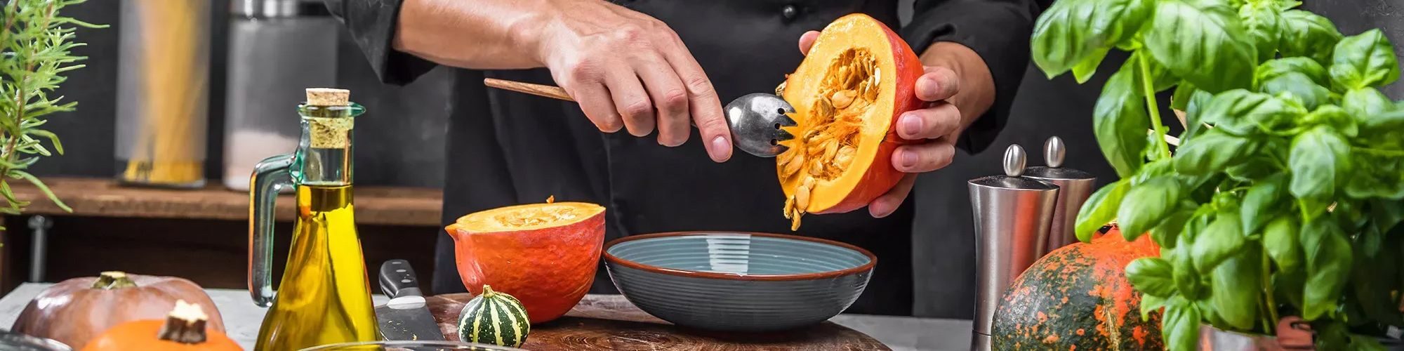 Image de restaurateur, dans une cuisine, entouré par des ingrédients : huile d'olive, et différentes courges. Il vide une courge dans un bol.