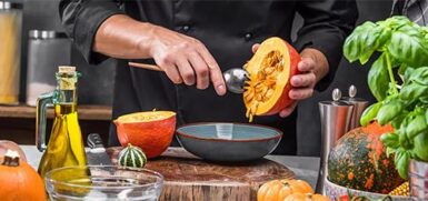Image de restaurateur, dans une cuisine, entouré par des ingrédients : huile d'olive, et différentes courges. Il vide une courge dans un bol.