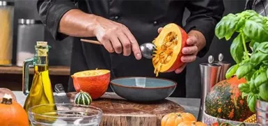 Image de restaurateur, dans une cuisine, entouré par des ingrédients : huile d'olive, et différentes courges. Il vide une courge dans un bol.