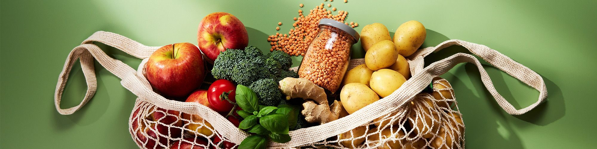 Un sac de courses qui déborde sur un fond vert. Dans le sac il y a des pommes, des tomates, des brocolis, des patates, des poids chiches, de la coriandre, etc