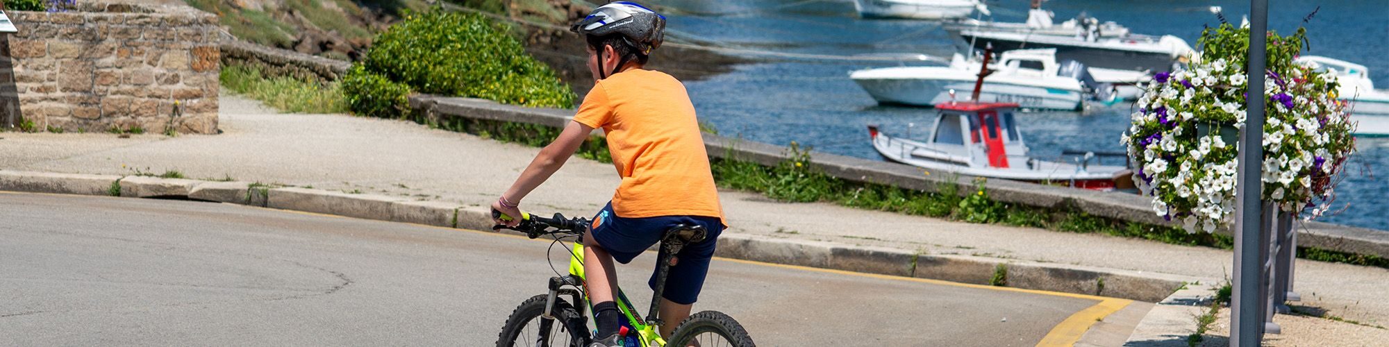 Un enfant qui roule à vélo sur la route, en arrière plan on peut voir le port et des bateaux.